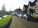 Image: Left of a road is a flat grass bank and the River Sheppey; to the rightm cottages face over the river