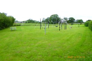 A wide open space, bordered by hedges with metal play equipment to the foreground and an open space for ball sports etc.