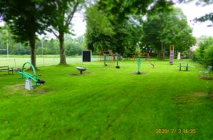 picture shows the fenced off play area, below well established trees in full leaf and a variety of play equipment