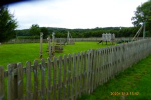 Picture shows a largely wooden activity playground, surrounded by a fence toward to top of a slope, next to a cricket pitch (Not owned by the Parish Council)